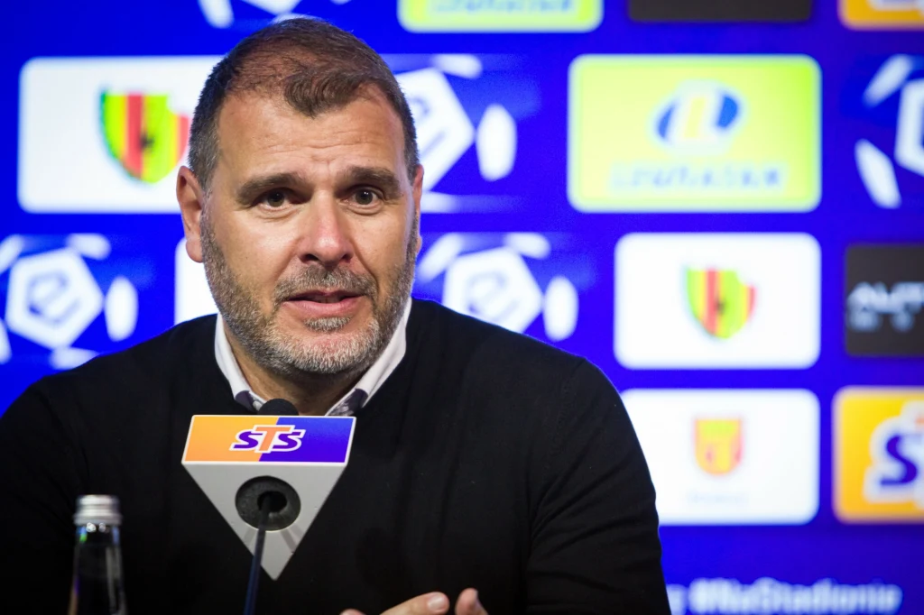 Mężczyzna z zarostem w ciemnym swetrze siedzi przy mikrofonie na tle niebieskiej ściany z logotypami sponsorów i emblematów sportowych.