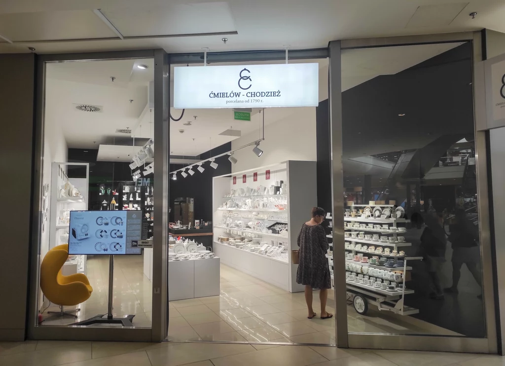 Witryna sklepu z porcelaną, półki wypełnione białą zastawą i akcesoriami, przy regale stoi kobieta oglądająca produkty, wnętrze jasne i nowoczesne.