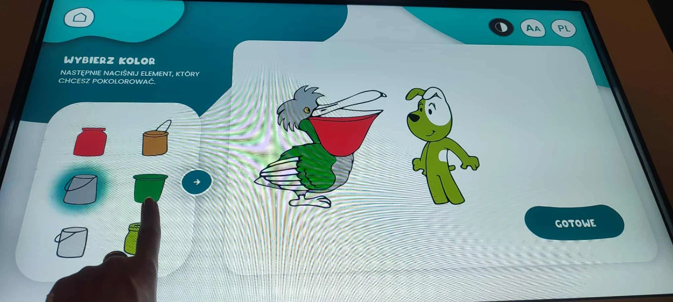 W Studio każdy może wyreżyserować swoją bajkę Fot./GabrielaWójcik Ekran dotykowy z aplikacją do kolorowania cyfrowych postaci bajkowych, pelikan trzyma wiadro oraz zielony pies, poniżej palec wskazuje wybór zielonego koloru farby.