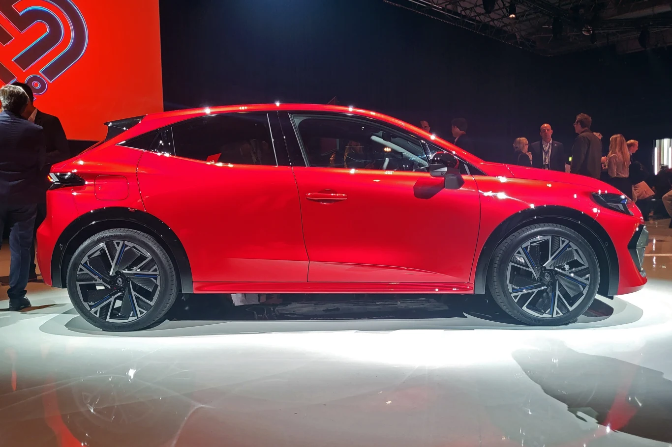 Nowoczesny czerwony samochód osobowy o sportowej sylwetce, ustawiony na błyszczącej podłodze, podświetlony w warunkach ekspozycyjnych, w tle kilku ludzi i elementy scenografii targowej.