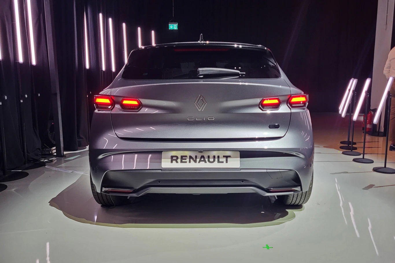 Srebrny samochód Renault Clio ustawiony tyłem na błyszczącej podłodze, otoczony pionowymi światłami LED, w tle elementy ekspozycji i słupki ochronne.