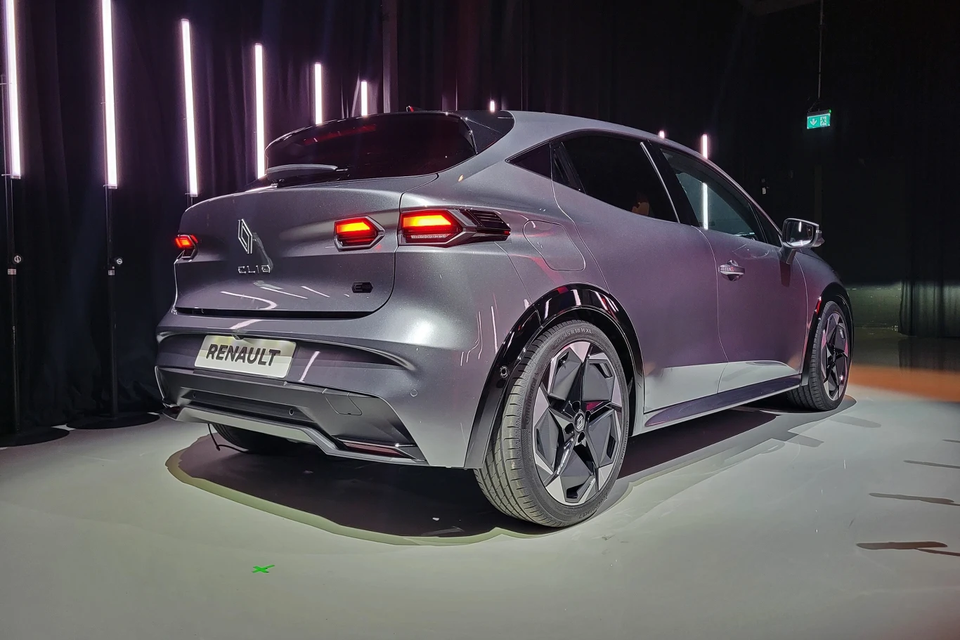 Srebrny samochód marki Renault o nowoczesnym, opływowym kształcie stoi na ekspozycji pod sztucznym oświetleniem. Widoczna jest tylna część pojazdu z charakterystycznymi reflektorami i oznaczeniem modelu na klapie bagażnika.