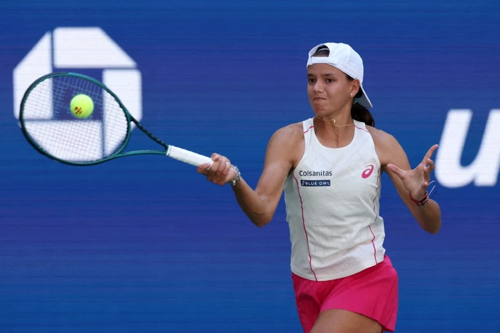 Tenisistka w białej czapce i sportowym stroju odbijająca piłkę podczas meczu na profesjonalnym korcie, z widocznym skupieniem na twarzy.