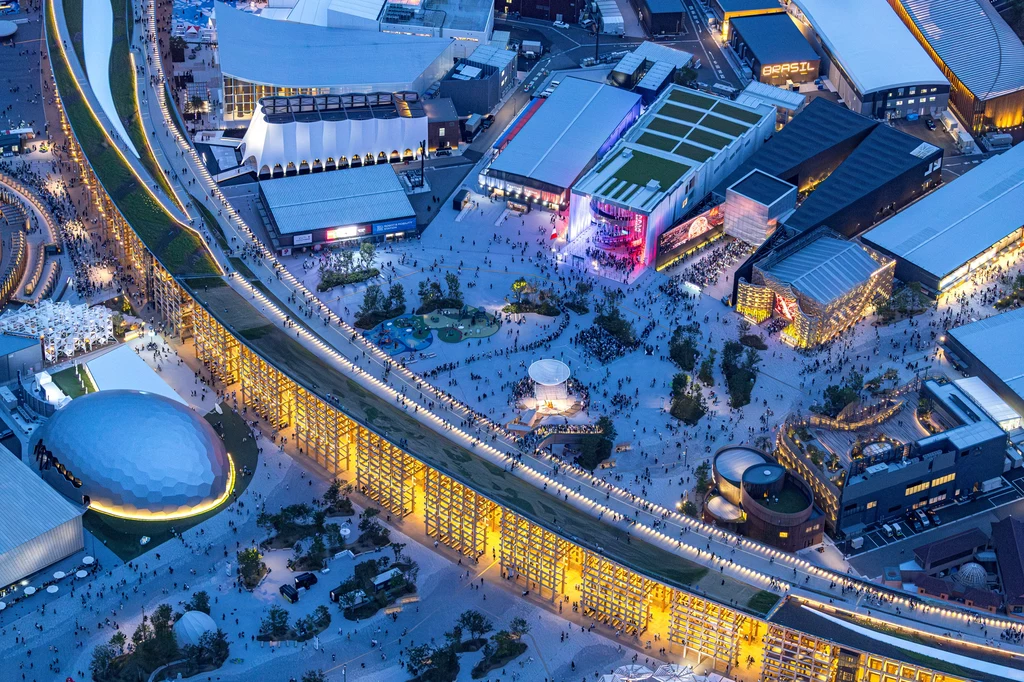 Duże skupisko ludzi poruszających się po terenach miejskich, otoczone nowoczesnymi budynkami i halami, dobrze oświetlona przestrzeń z różnorodną architekturą oraz dominantą w postaci długiego budynku z licznymi światłami.