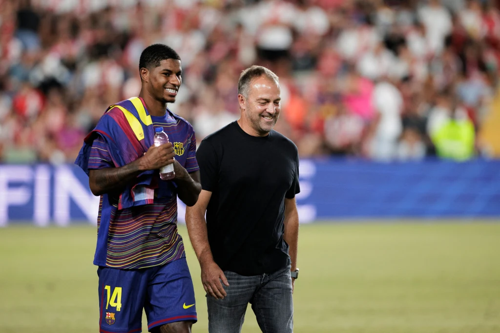 Marcus Rashford i Hansi Flick Uśmiechnięty piłkarz w kolorowej koszulce FC Barcelona idzie obok dorosłego mężczyzny ubrany w czarną koszulkę i dżinsy, otoczony rozmytym tłumem kibiców na stadionie piłkarskim.