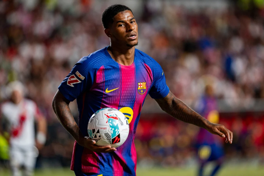 Marcus Rashford Piłkarz w stroju FC Barcelony trzyma piłkę pod pachą podczas meczu, w tle niewyraźni kibice na trybunach.