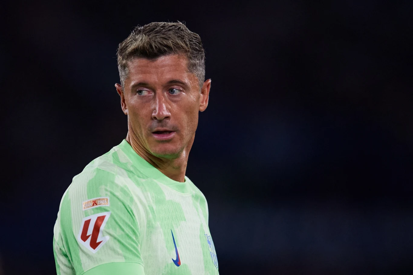 Mężczyzna w jasnozielonej sportowej koszulce z krótkimi rękawami, patrzący przez ramię, ciemne tło podkreśla jego sylwetkę.