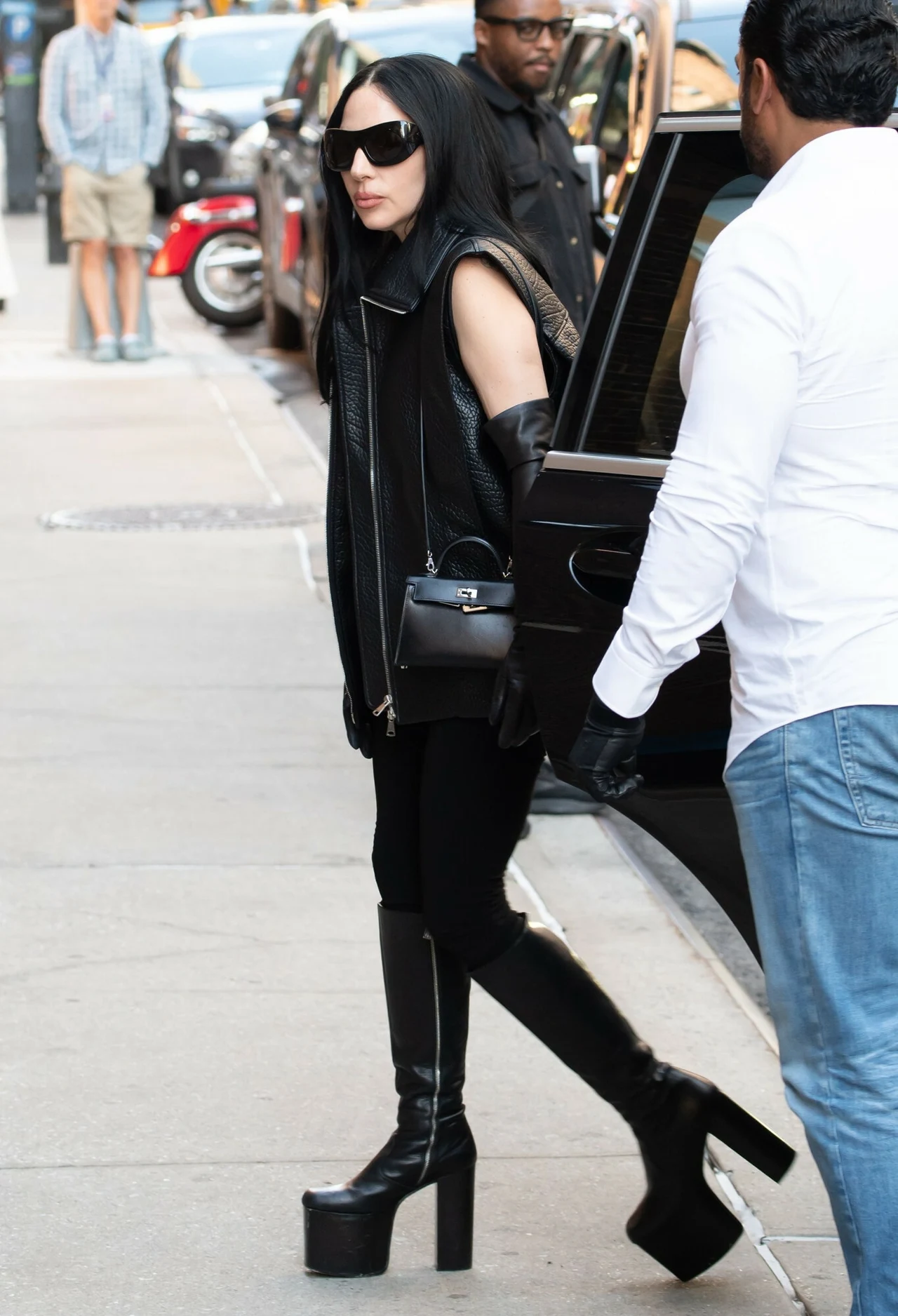 Kobieta w czarnych ubraniach i wysokich platformach wychodzi z czarnego samochodu, na twarzy czarne okulary przeciwsłoneczne oraz czarne rękawiczki, trzyma czarną torebkę, obok stoi mężczyzna w białej koszuli, w tle piesi na chodniku.