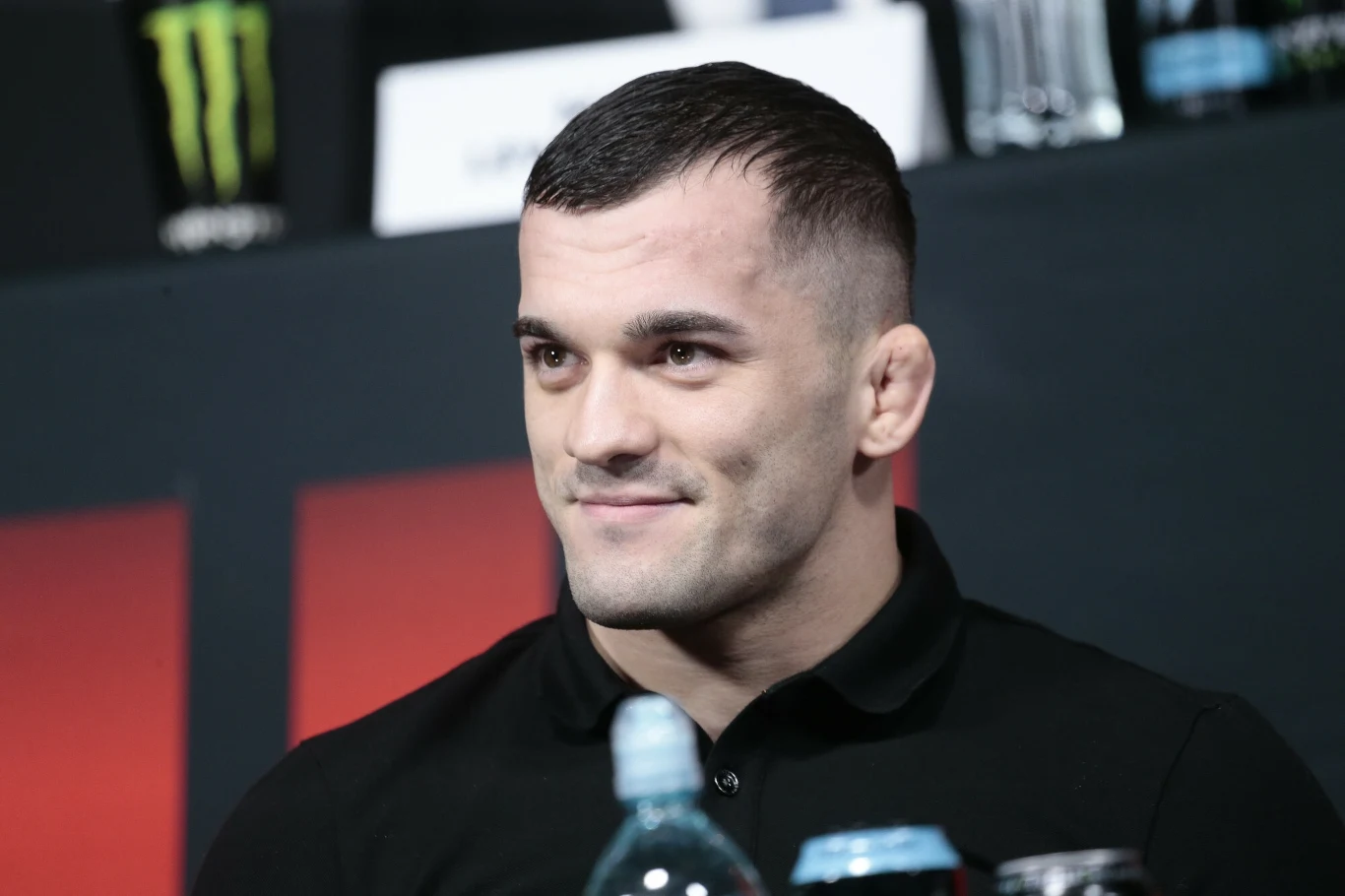 Mężczyzna o krótkich włosach i ciemnej koszulce siedzący przy stole podczas konferencji prasowej, w tle widoczne butelki z napojami i materiały promocyjne.