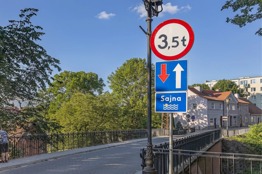 Most z żelazną barierką prowadzący nad wodą, przy którym znajdują się znaki drogowe ograniczające dopuszczalny tonaż pojazdów oraz regulujące pierwszeństwo przejazdu, w tle domy oraz zieleń drzew.