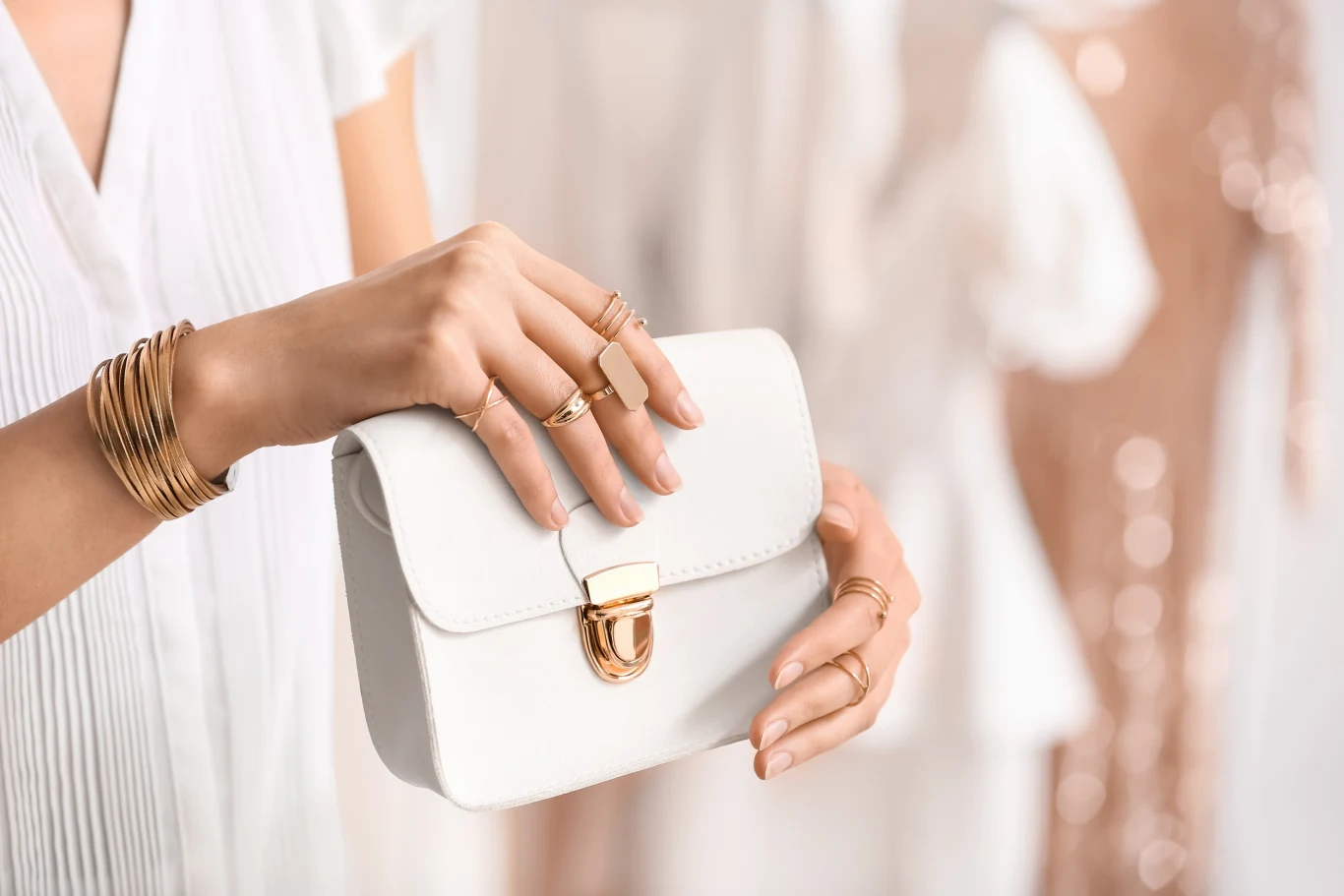 Sposób noszenia pierścionków ma wpływ na nasze finanse? Damska dłoń trzymająca elegancką, białą kopertówkę z metalowym zapięciem, ozdobiona licznymi pierścionkami i bransoletkami, w tle rozmyte sylwetki jasnych sukienek.