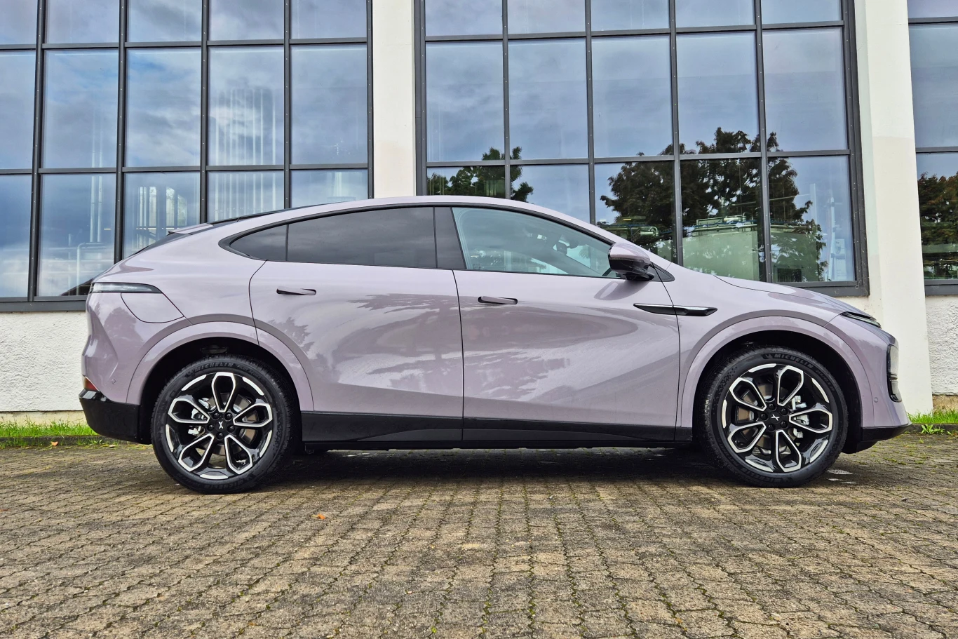 Nowy lakier Stellar Purple to kolejny wyróżnik odświeżonego Xpenga G6 Samochód osobowy o nowoczesnej, opływowej sylwetce ustawiony bokiem na tle dużych okien, pojazd stoi na brukowanej powierzchni.