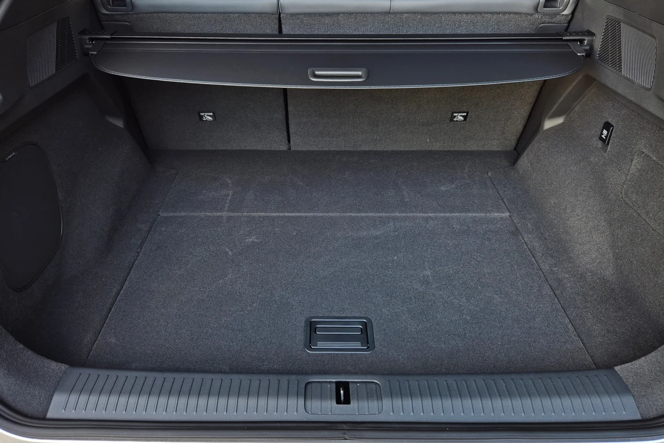 Bagażnik w Xpengu G6 ma 571 l, ale zaskakująco nisko umieszczona roleta wyraźnie zmniejsza możliwości przewozowe Pusta, czysta przestrzeń bagażnika samochodu z rozłożoną roletą ochronną, wnętrze wykończone ciemną tapicerką materiałową, widoczne zaczepy oraz schowek w podłodze.