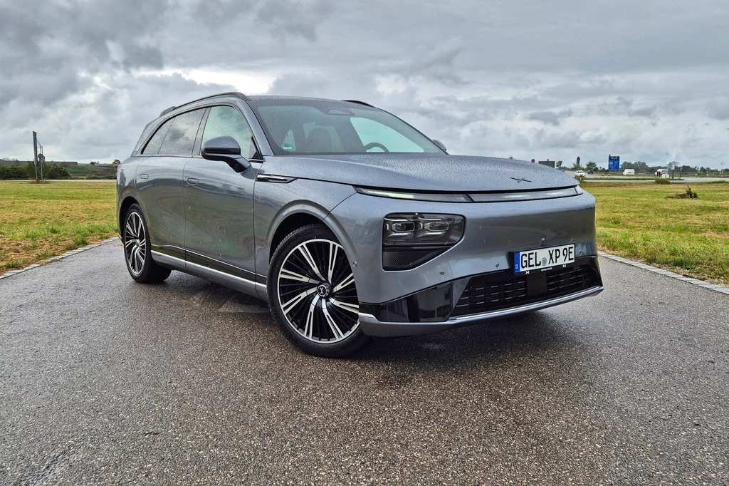 Xpeng G9 po face liftingu wygląda niemal identycznie co poprzednik Srebrny samochód elektryczny typu SUV stoi na mokrym asfalcie, widoczny z przodu, z charakterystycznymi reflektorami LED, aluminiowymi felgami oraz niemiecką tablicą rejestracyjną, tło stanowi zielone pole i pochmurne niebo.