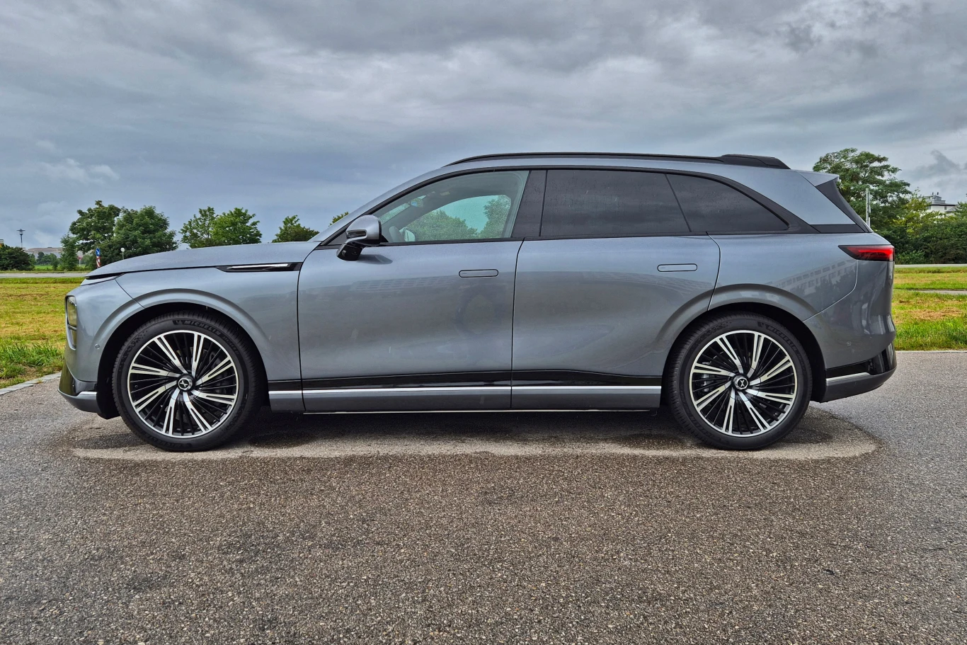 W najmocniejszej wersji Xpeng G9 wyposażony jest w pneumatyczne zawieszenie z regulacją prześwitu oraz siły tłumienia nierówności Srebrny samochód typu SUV zaparkowany na asfaltowej nawierzchni, po bokach rozciąga się zielona łąka i pojedyncze drzewa, niebo zachmurzone.