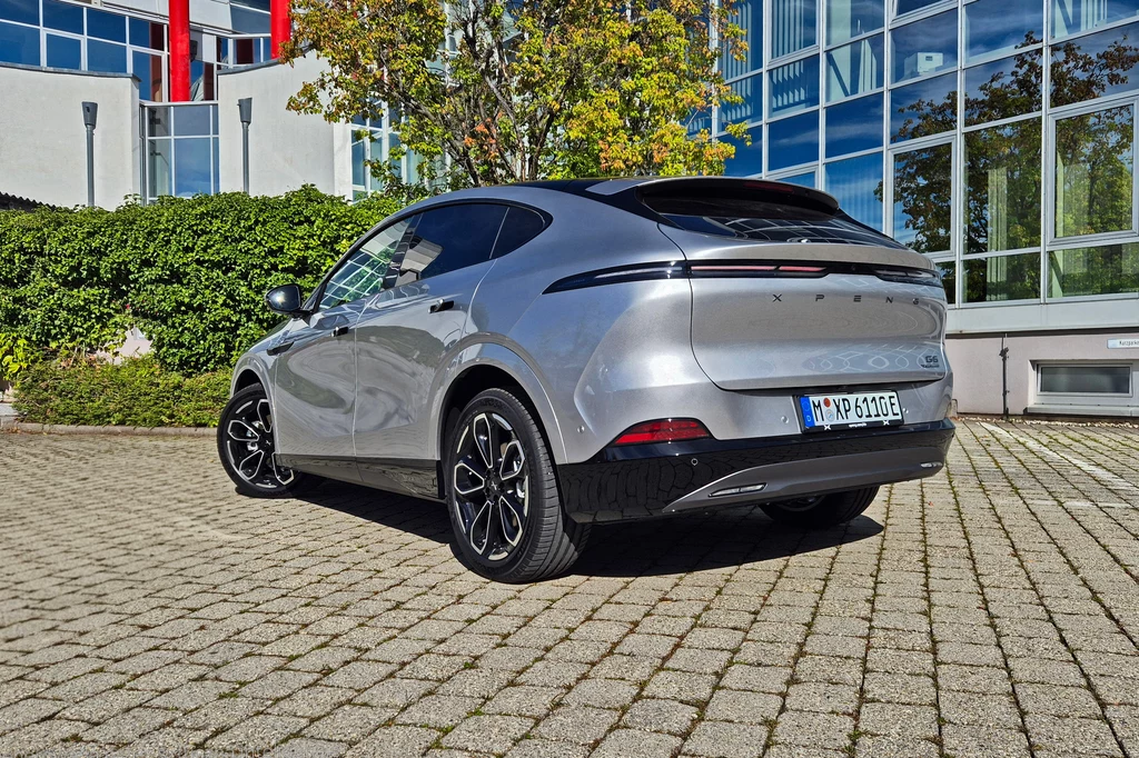 Odświeżony Xpeng G6 otrzymał zmieniony dyfuzor, a zintegrowany spojler na klapie bagażnika jest nieznacznie większy Nowoczesny srebrny samochód elektryczny typu SUV zaparkowany na brukowanym placu, otoczony nowoczesnymi szklanymi budynkami oraz zielenią.