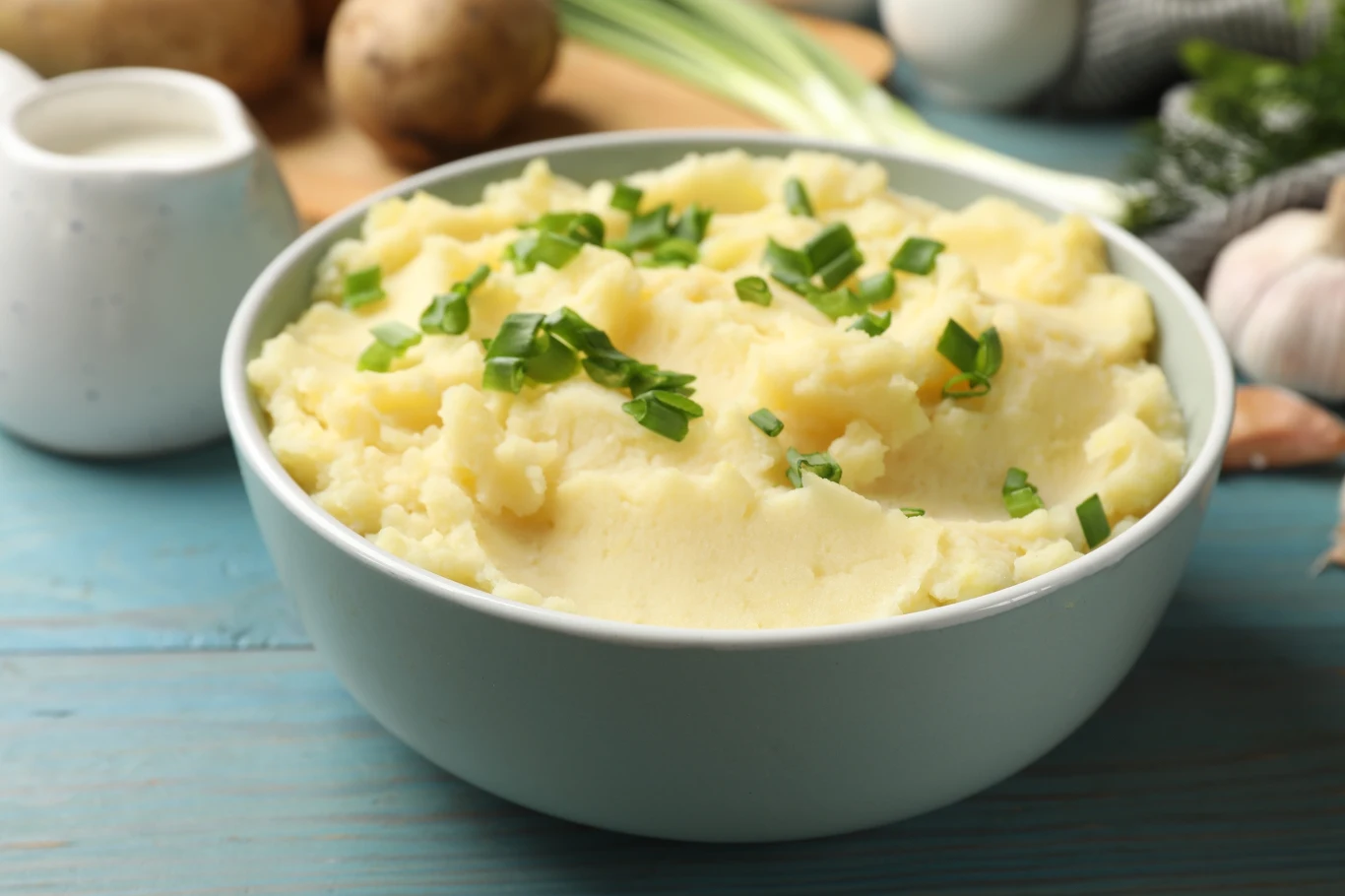 Idealne w smaku i konsystencji puree ziemniaczane to prawdziwy przysmak w polskiej kuchni Porcja kremowego puree ziemniaczanego w misce, udekorowana świeżymi drobno posiekanymi szczypiorkiem, w tle warzywa i dzbanek ze śmietaną.