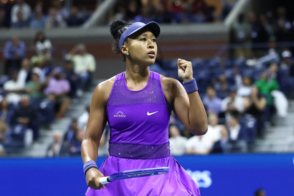 Naomi Osaka Tenisistka ubrana w fioletowy strój na korcie, z uniesioną pięścią w geście zwycięstwa, na tle widowni z rozmytą publicznością.