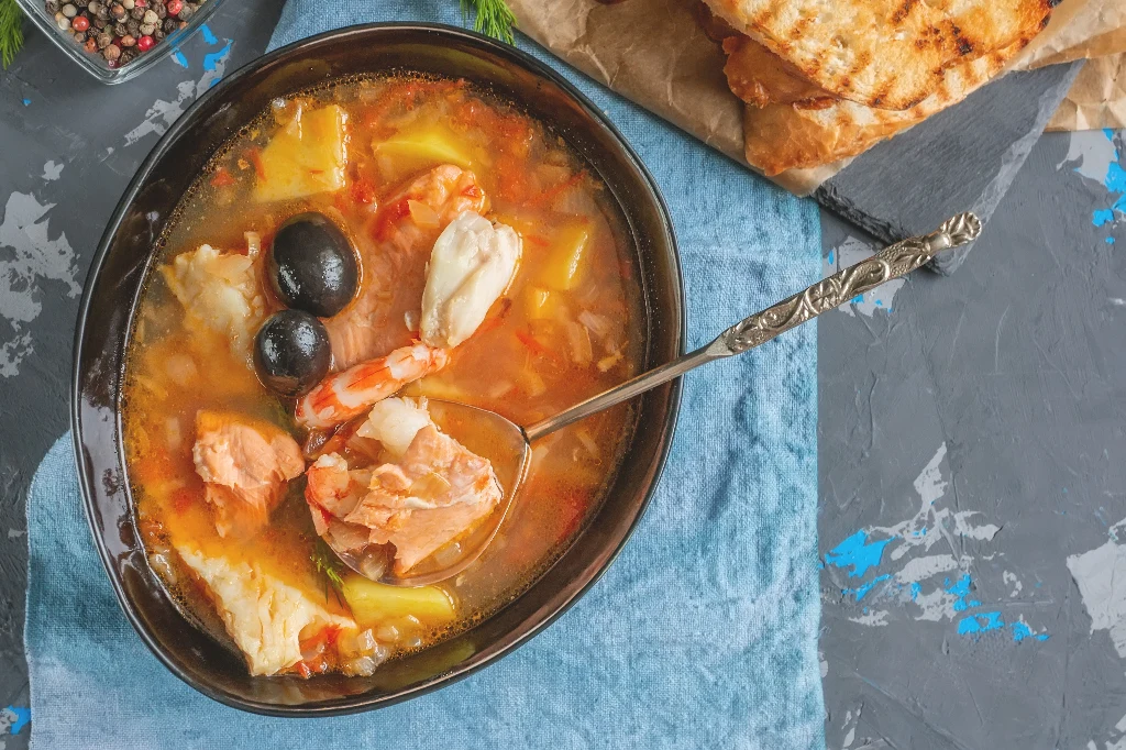 Zupa rybna z kawałkami ryby, krewetką, oliwkami i ziemniakami w jasnym bulionie, podawana w głębokim talerzu na niebieskiej serwetce, obok grillowane pieczywo.