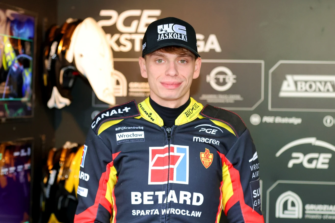 Młody mężczyzna w stroju żużlowca, z logo sponsorów i nazwą klubu BETARD Sparta Wrocław, stoi na tle ścianki promocyjnej z logotypami ligi oraz partnerów.