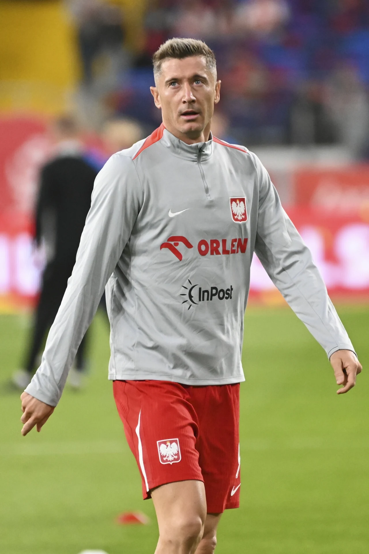 Piłkarz w szarej bluzie treningowej reprezentacji Polski oraz czerwonych spodenkach stoi na boisku podczas rozgrzewki, w tle zamazane postaci oraz elementy stadionu.