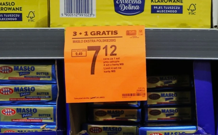 Pomarańczowa etykieta promocyjna informująca o ofercie 3+1 gratis na masło ekstra polskie 200 g, ułożone opakowania masła na półce sklepowej, wokół inne produkty mleczne.