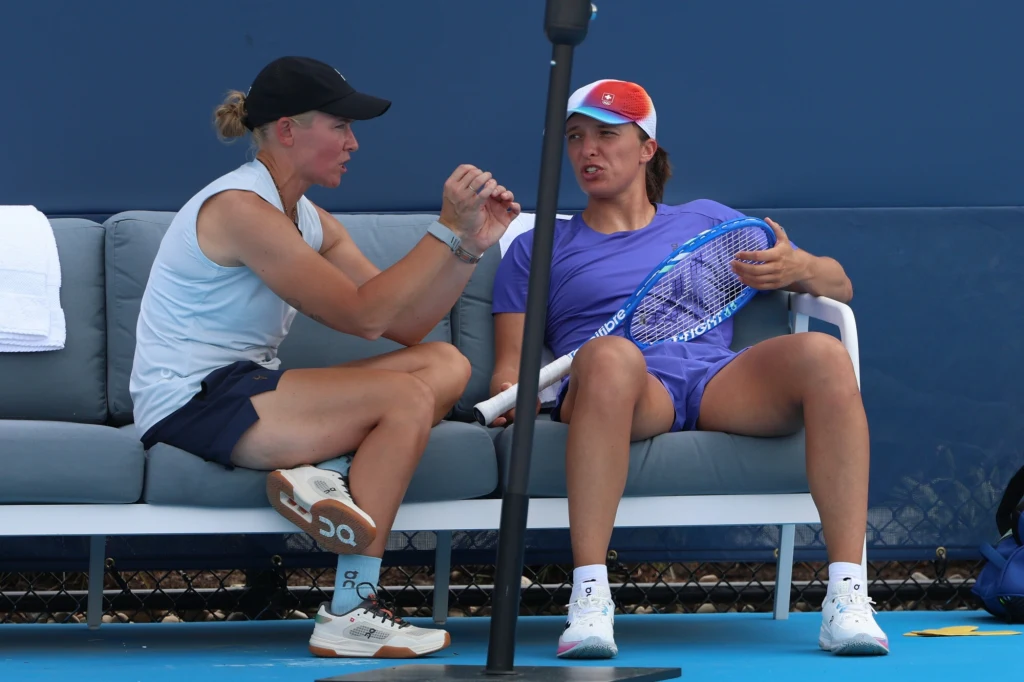 Daria Abramowicz i Iga Świątek Dwie kobiety siedzą na ławce sportowej, jedna trzyma rakietę tenisową i ubrane są w sportowe stroje, rozmawiają ze sobą w trakcie odpoczynku na otwartym korcie tenisowym.