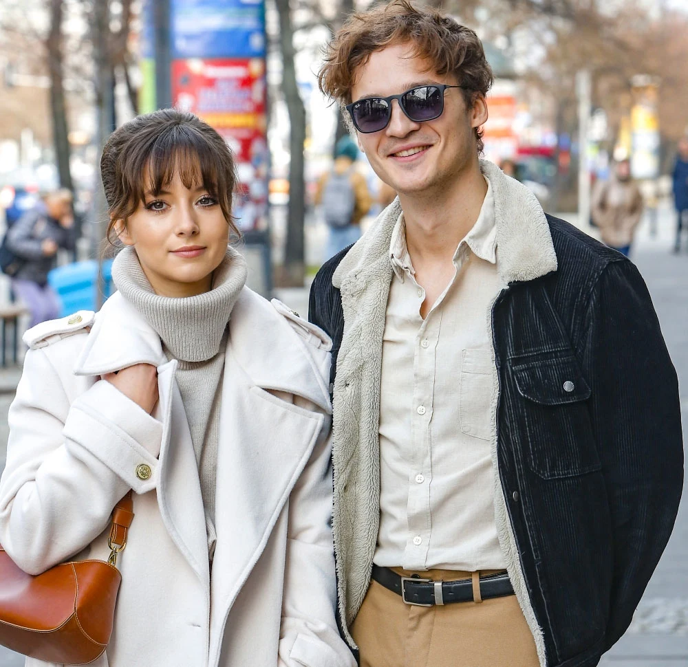 Kasia i Jacek Sienkiewiczowie robią sobie przerwę od wspólnych działań pod szyldem Kwiat Jabłoni Młoda kobieta w jasnym płaszczu i golfie stoi obok uśmiechniętego mężczyzny w ciemnej kurtce z kożuchem, jasnej koszuli i okularach przeciwsłonecznych, para na tle ulicy z drzewami i przechodniami, w tle rozmyte kolorowe plakaty reklamowe.