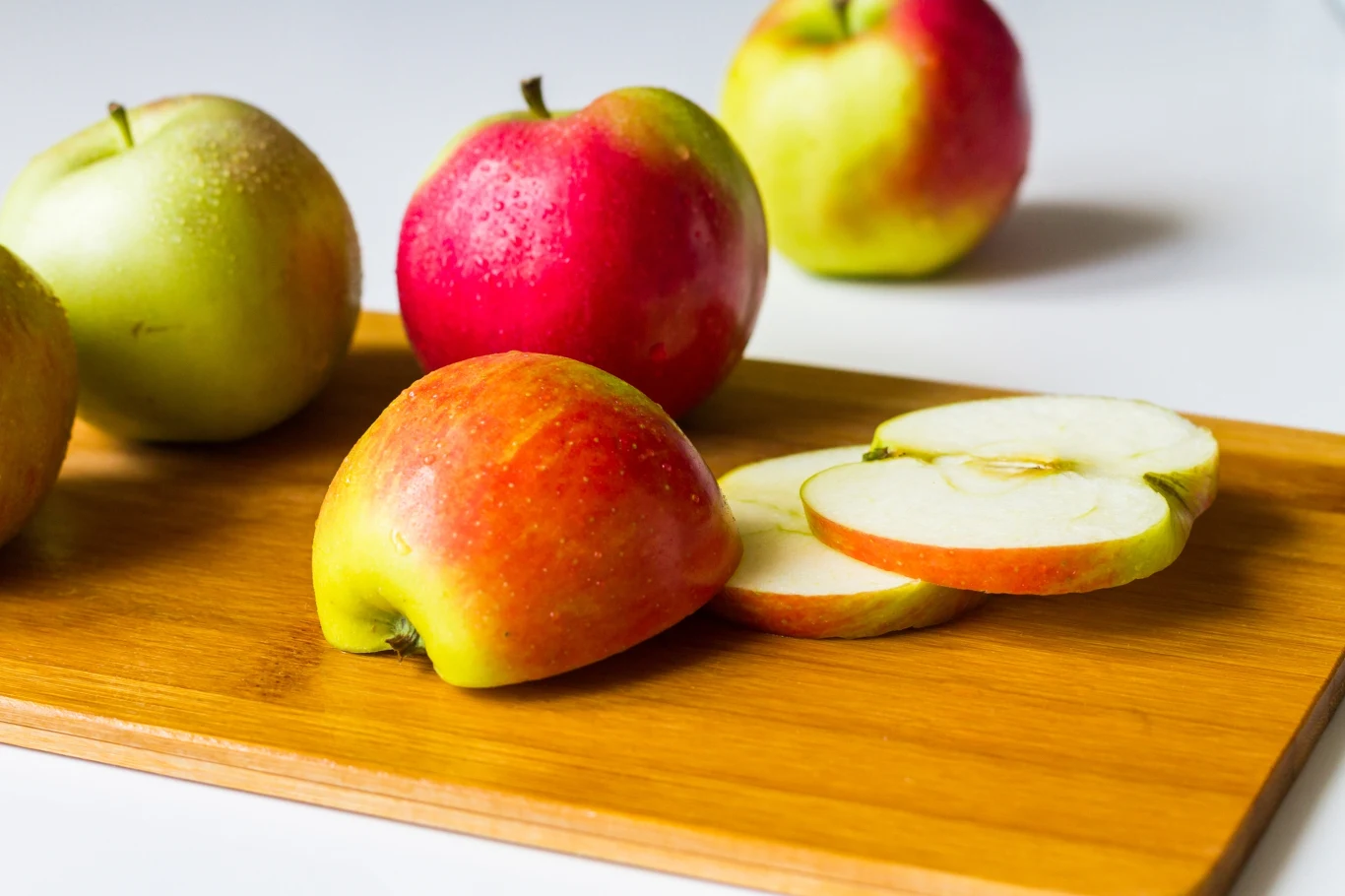 Kilka kolorowych jabłek leżących na drewnianej desce, jedno z nich pokrojone w plastry, reszta w całości; tło rozmyte, jasne.