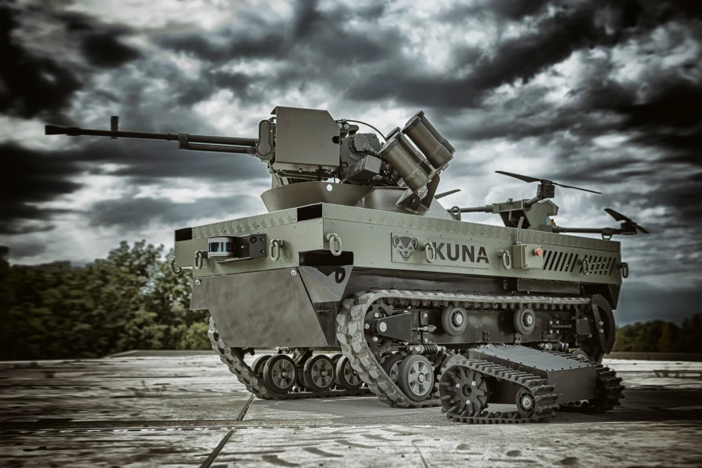 Nowoczesny, bezzałogowy pojazd gąsienicowy o militarnej konstrukcji, wyposażony w różne typy uzbrojenia i czujniki. Maszyna stoi na betonowej powierzchni, na tle burzowego nieba i drzew w oddali.