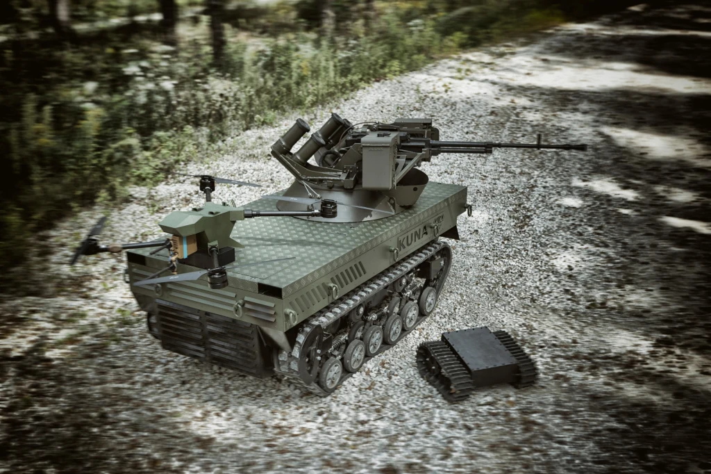 Zdalnie sterowany pojazd wojskowy na gąsienicach wyposażony w zamontowane uzbrojenie, w otoczeniu leśnym na żwirowej drodze, obok mniejszy pojazd mobilny również z napędem gąsienicowym.