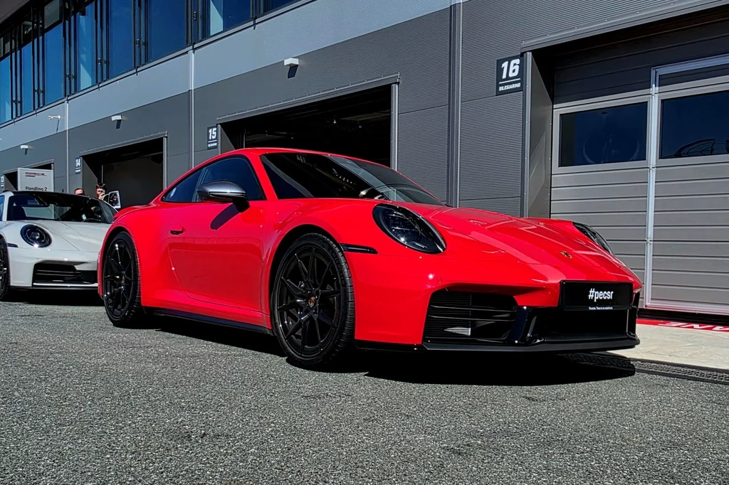Czerwony sportowy samochód Porsche zaparkowany przed nowoczesnym budynkiem, w tle widoczny biały samochód oraz szare drzwi garażowe z numeracją.