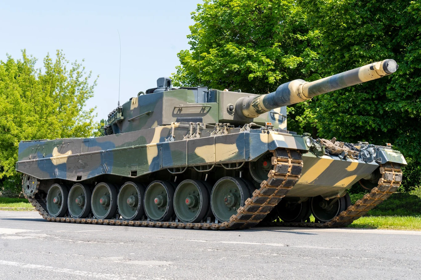 Czołg Leopard 2 w wersji z kamuflażem, stojący na asfalcie, otoczony zielenią drzew, widoczny bok i lufa skierowana w lewo, pojazd w bardzo dobrym stanie technicznym.