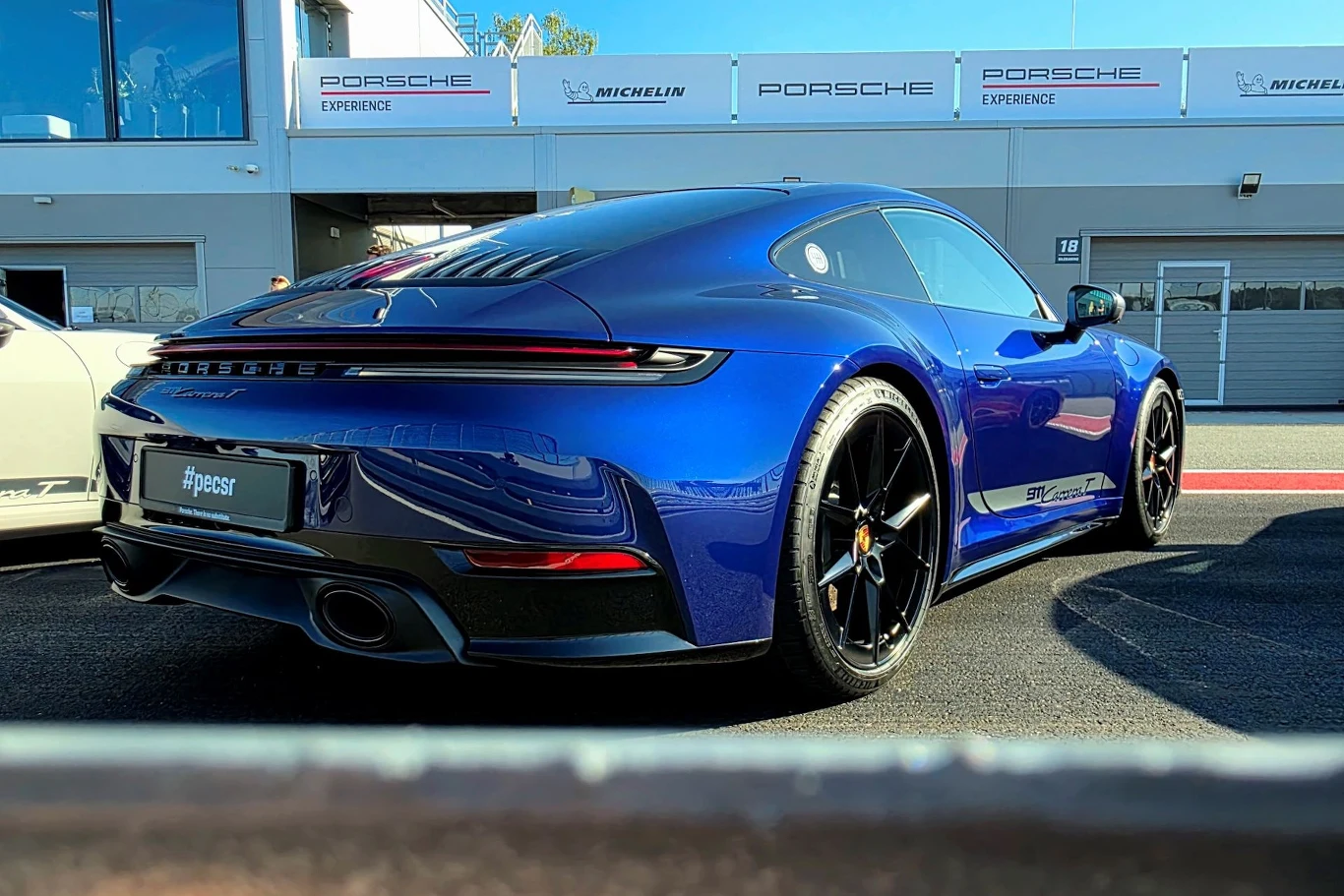 Niebieski samochód sportowy marki Porsche z czarnymi felgami stoi na asfaltowej nawierzchni przed nowoczesnym budynkiem, na którym widoczne są logotypy Porsche Experience i Michelin.