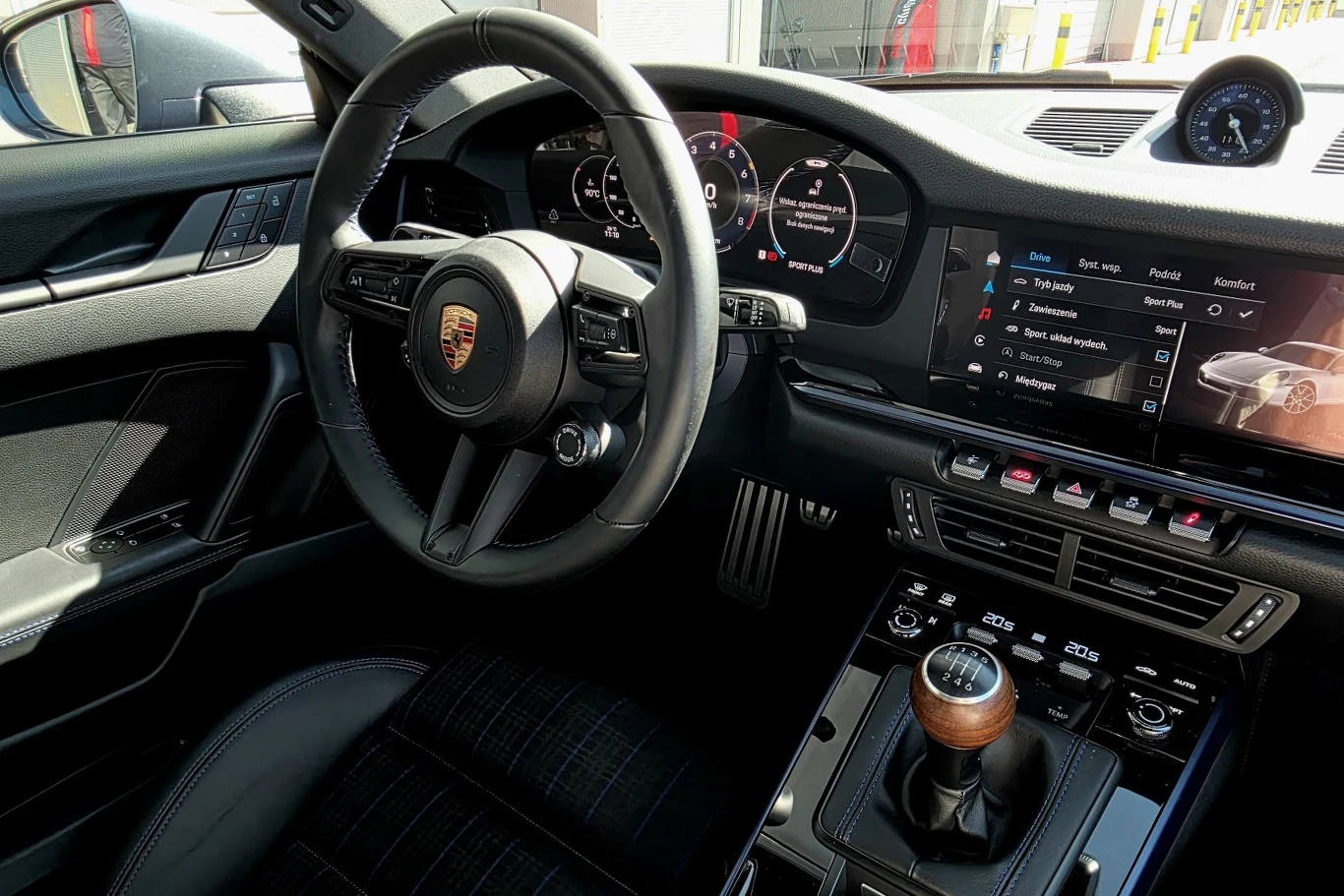 Luksusowe wnętrze samochodu marki Porsche z widocznym logotypem na kierownicy, manualną skrzynią biegów z drewnianą gałką, cyfrowymi zegarami na desce rozdzielczej oraz nowoczesnym systemem multimedialnym.