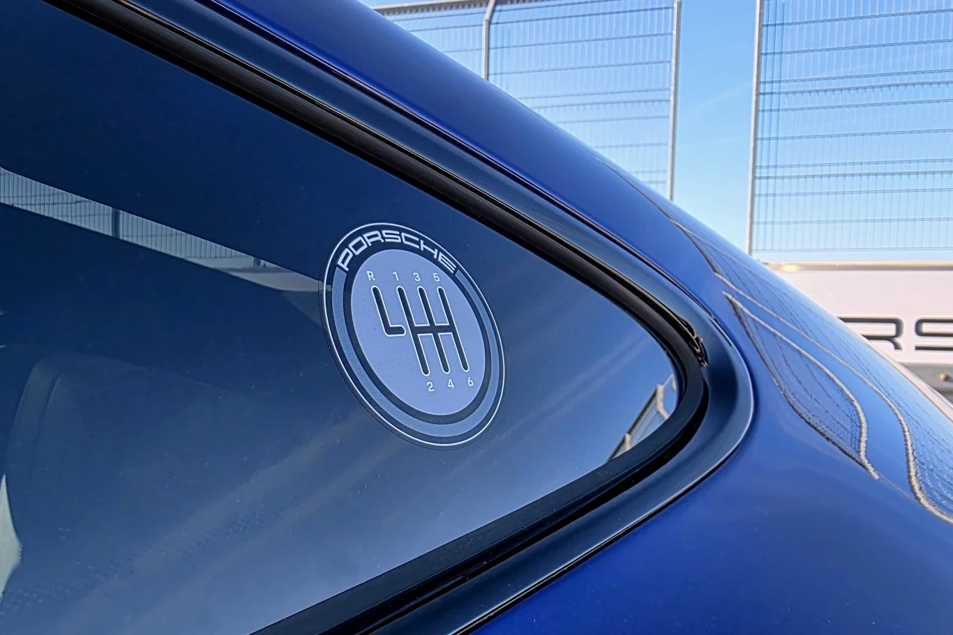 Szyba boczna niebieskiego samochodu z widoczną naklejką Porsche z motywem schematu manualnej skrzyni biegów, w tle stalowe ogrodzenie i fragment tablicy informacyjnej.