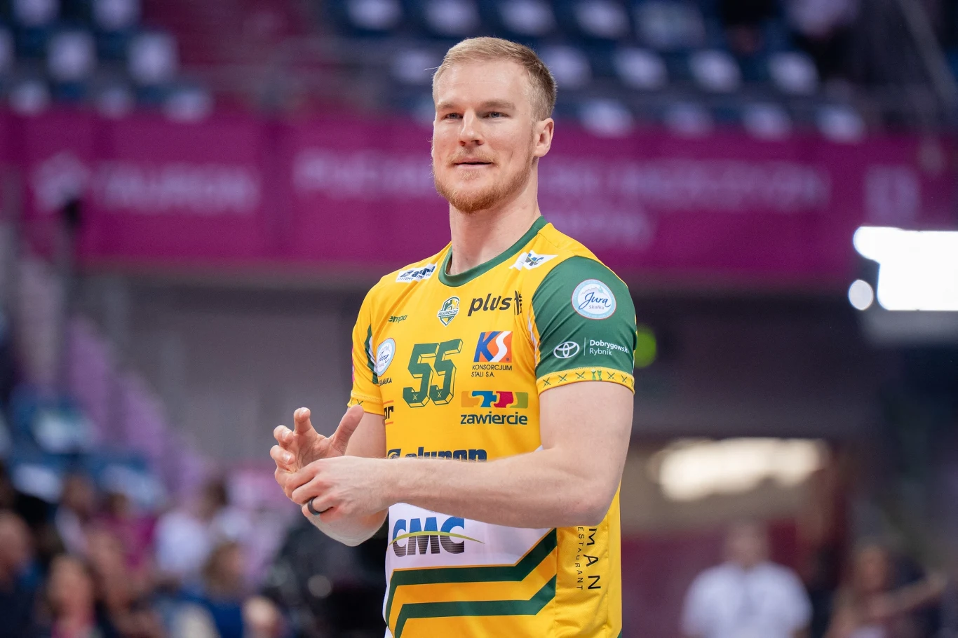 Siatkarz w żółto-zielonym stroju sportowym z numerem 55 stoi na boisku, w tle widoczne niewyraźne trybuny i kibice.