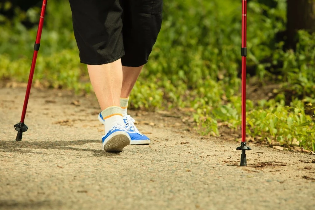 Nogi osoby idącej po leśnej ścieżce z kijkami do nordic walking, sportowe buty i zielone otoczenie lasu w tle.