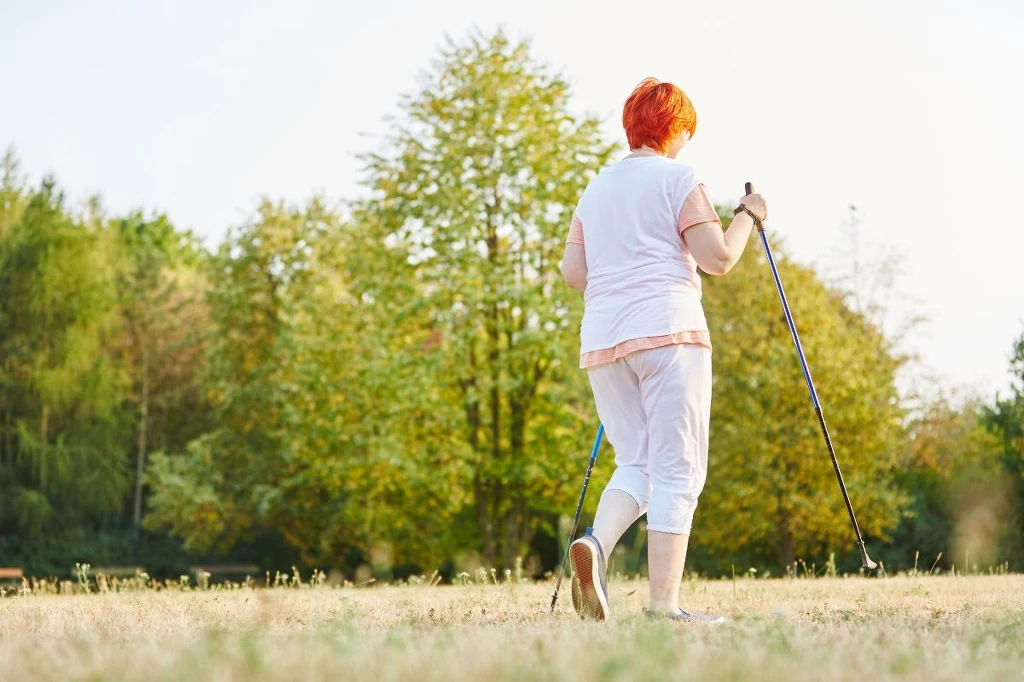 Kobieta z rudymi włosami spaceruje z kijkami nordic walking po polanie otoczonej drzewami, ubrana w jasne, sportowe ubranie, słoneczna pogoda i zielone otoczenie wskazują na aktywny wypoczynek na świeżym powietrzu.
