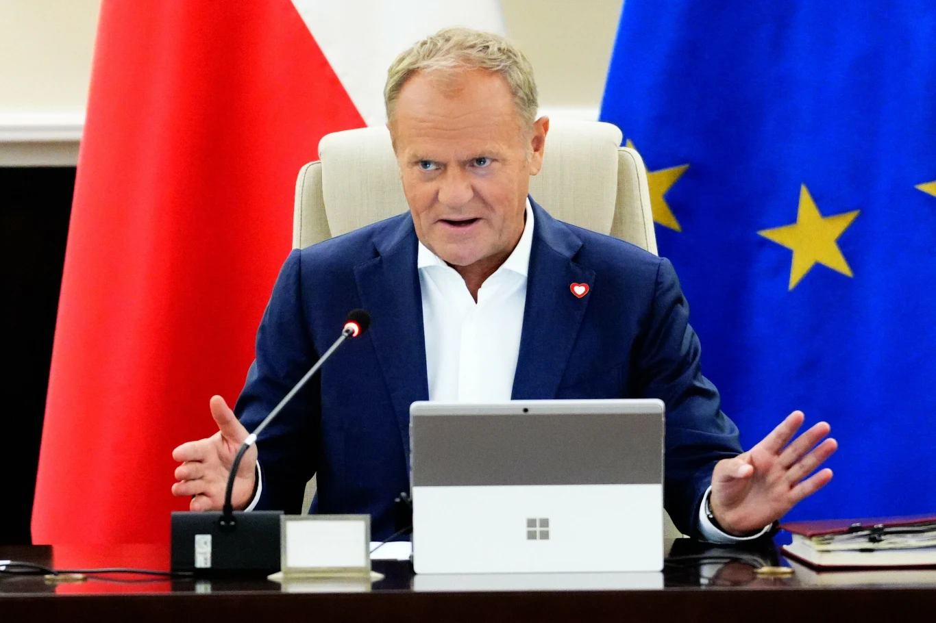 Premier Donald Tusk zapowiada nowy projekt ustawy dotyczący zasad wypłacania świadczenia 800 plus Ukraińcom w Polsce Mężczyzna w garniturze siedzący przy stole konferencyjnym, mówiący energicznie, przed nim laptop, mikrofon i dokumenty, w tle widoczne flagi Polski oraz Unii Europejskiej.