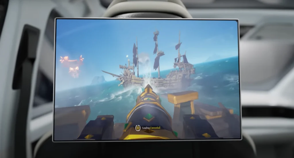 Xbox Ekran monitora w samochodzie wyświetla scenę z gry komputerowej o tematyce pirackiej, ukazując widok z perspektywy działa okrętowego skierowanego w stronę statku na morzu z rozpostartymi czarnymi żaglami.
