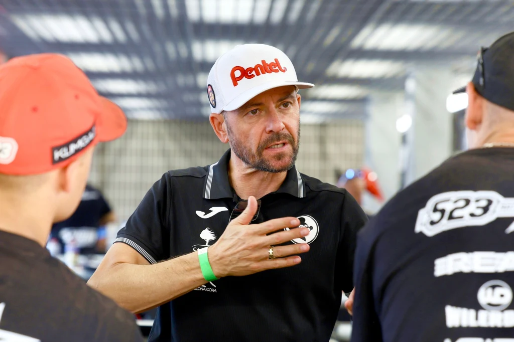 Piotr Protasiewicz, legenda Falubazu. Mężczyzna w czapce z daszkiem i koszulce sportowej gestykuluje podczas rozmowy z dwoma innymi osobami, znajduje się w jasnym, zadaszonym pomieszczeniu.