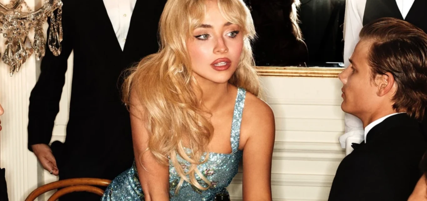 Kobieta w błyszczącej, niebieskiej sukni siedzi przy stole w towarzystwie elegancko ubranych osób, atmosfera wskazuje na przyjęcie lub formalne spotkanie.