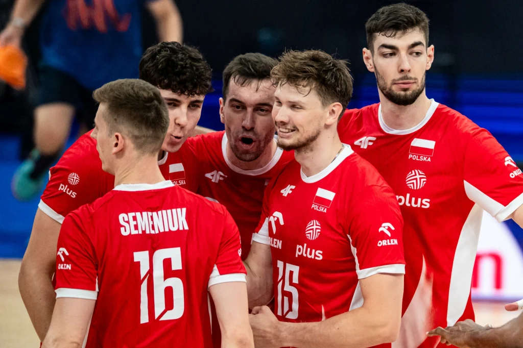 Grupa polskich siatkarzy w czerwonych strojach z widocznymi nazwiskami i numerami, świętujący wspólny sukces na boisku, wyrażający radość i zaangażowanie sportowe.