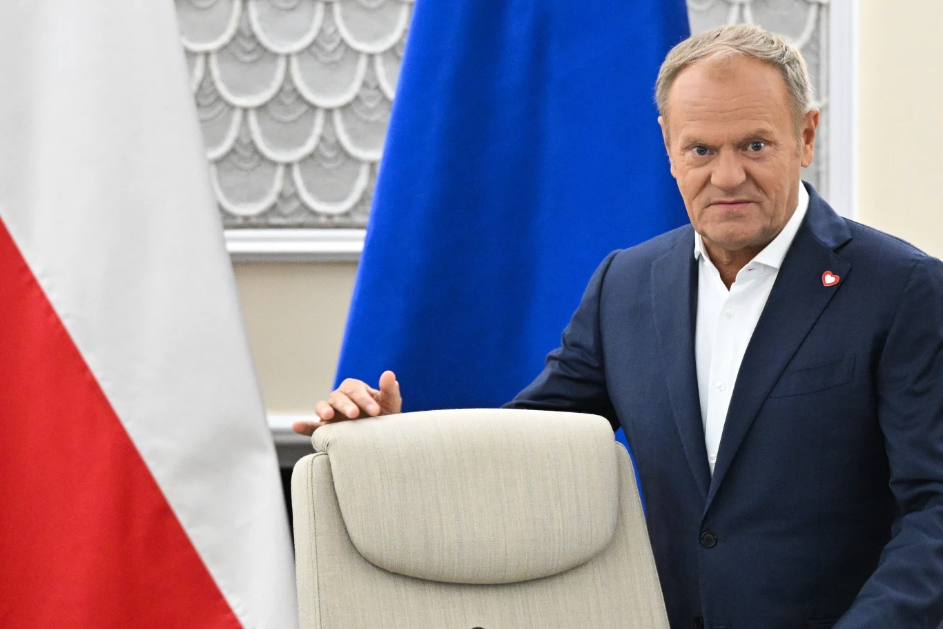 Donald Tusk podkreślił, że popiera kandydaturę Mariusza Haładyja na prezesa NIK Mężczyzna w garniturze stoi obok fotela na tle flagi Polski i niebieskiej flagi; w tle dekoracyjna ściana z elementami ozdobnymi.