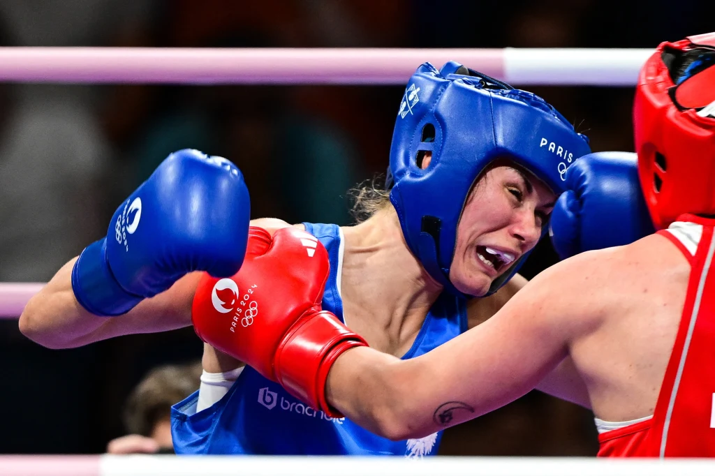 Aneta Rygielska Dwie zawodniczki w kaskach ochronnych i rękawicach bokserskich podczas walki w ringu, jedna z nich otrzymuje cios w twarz.