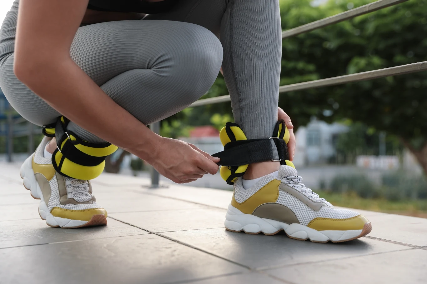 Osoba w sportowym stroju zapina obciążniki na kostkach, klęcząc na zewnątrz w pobliżu metalowej barierki, na tle zieleni.
