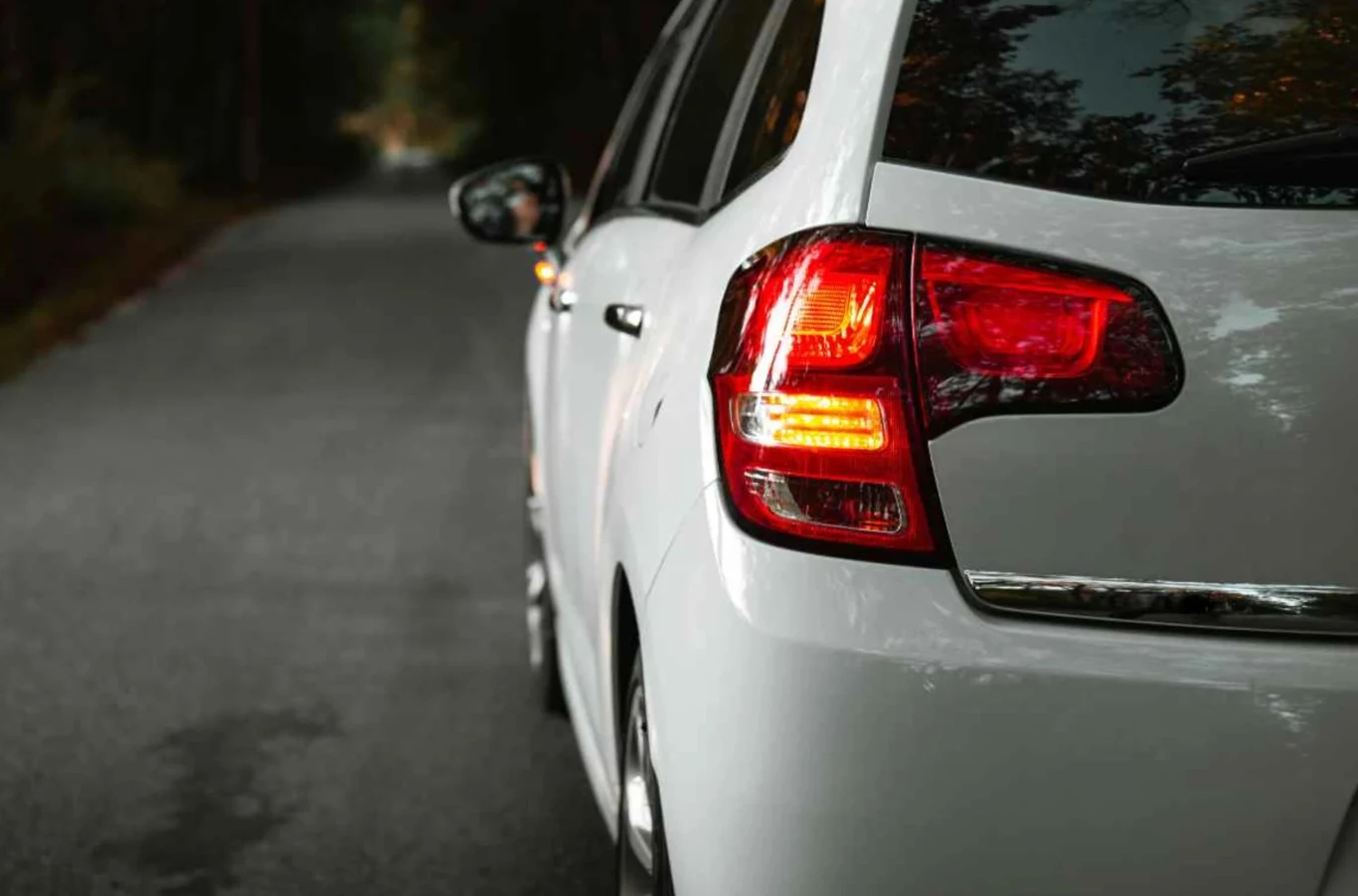 Czy można wyprzedzać auto skręcające w lewo? Biały samochód osobowy zaparkowany przy skraju asfaltowej drogi otoczonej drzewami, włączone tylne światła pozycyjne