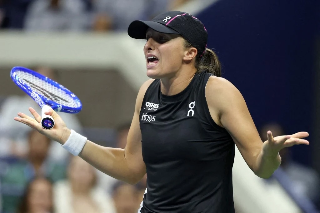 Iga Świątek Zawodniczka tenisa ubrana w czarny strój sportowy i czapkę z daszkiem wyraża emocje, trzymając rakietę tenisową i gestykulując dłońmi podczas meczu.