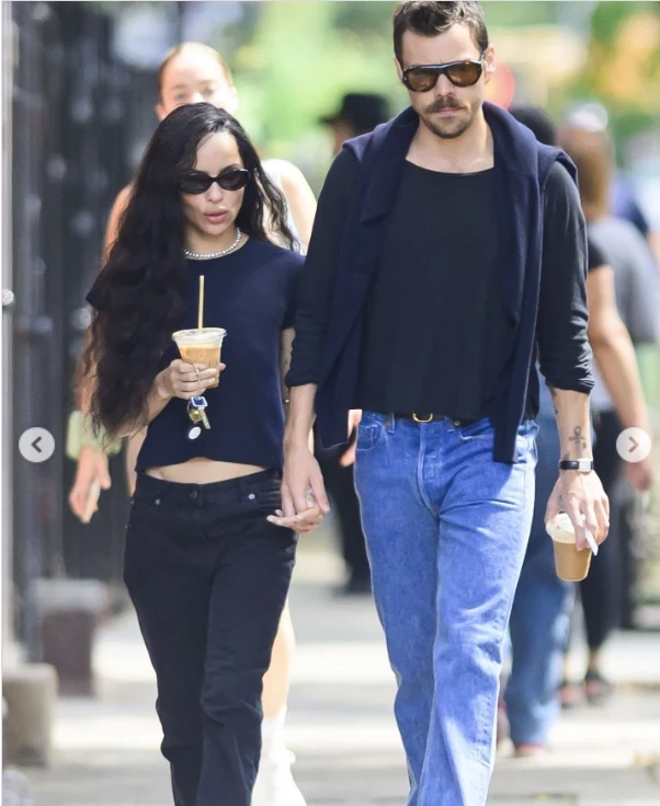 Para trzymająca się za ręce spaceruje po mieście, oboje ubrani w ciemne ubrania i noszący okulary przeciwsłoneczne, każde z nich trzyma kawę na wynos.